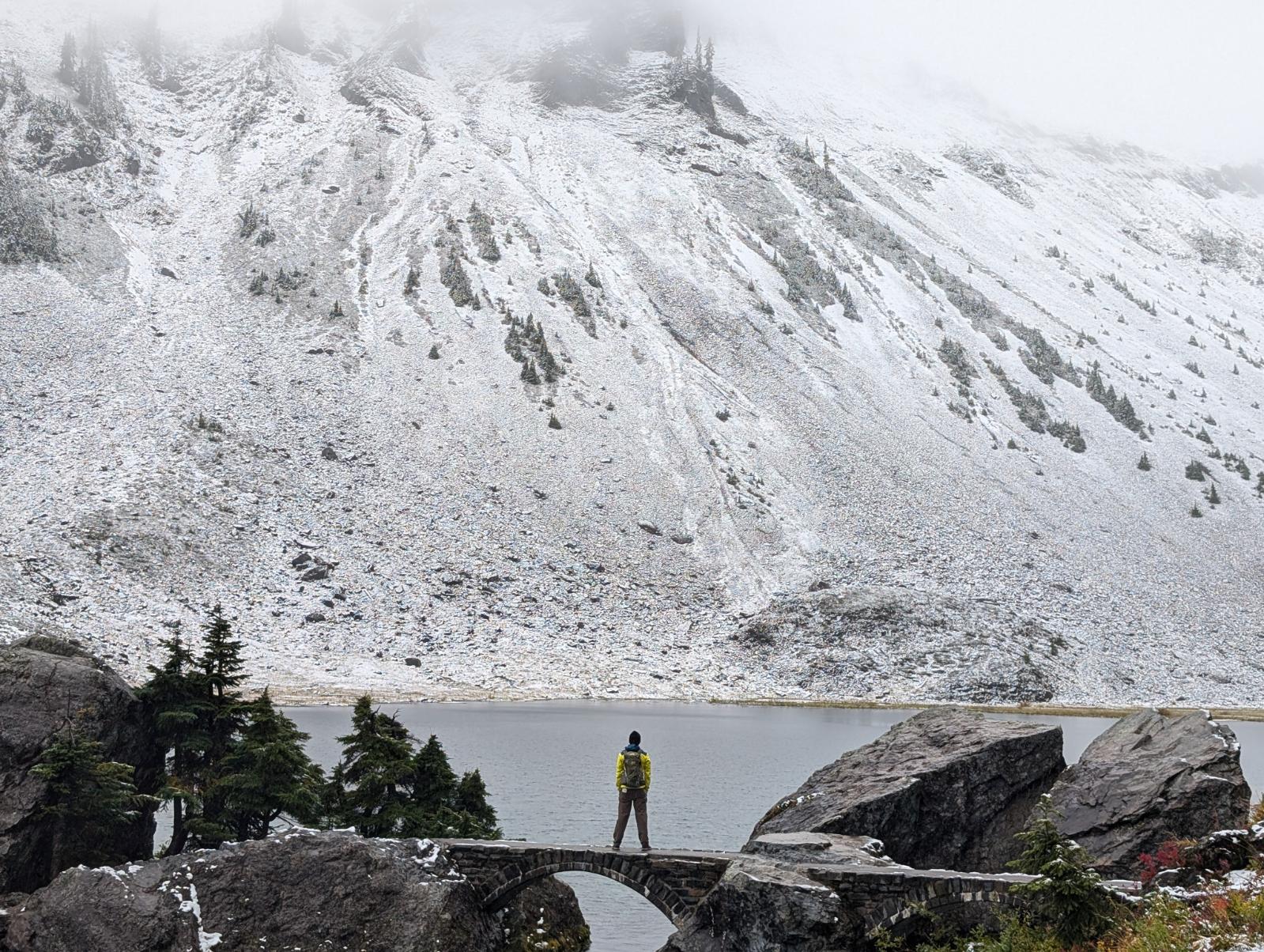John Hurr standing on a stone bridge overlooking a snowy mountain lake